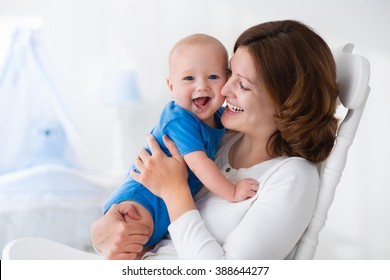 Young Mother Holding Her Newborn Child. Mom Nursing Baby. Woman And New Born Boy In White Bedroom With Rocking Chair And Blue Crib. Nursery Interior. Mother Playing With Laughing Kid. Family At Home