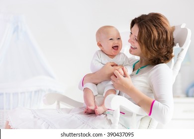 Young Mother Holding Her Newborn Child. Mom Nursing Baby. Woman And New Born Boy In White Bedroom With Rocking Chair And Blue Crib. Nursery Interior. Mother Playing With Laughing Kid. Family At Home