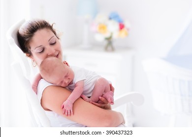 Young Mother Holding Her Newborn Child. Mom Nursing Baby. Woman And New Born Boy Relax In A White Bedroom With Rocking Chair And Blue Crib. Nursery Interior. Mother Breast Feeding Baby. Family At Home