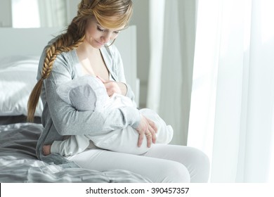 Young Mother Holding Her Newborn Child. Mom Nursing Baby. Woman And New Born Boy Relax In A White Bedroom. Mother Breast Feeding Baby. Family At Home