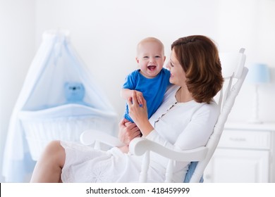 Young Mother Holding Her Newborn Child. Mom Nursing Baby. Woman And New Born Boy In White Bedroom With Rocking Chair And Blue Crib. Nursery Interior. Mother Playing With Laughing Kid. Family At Home