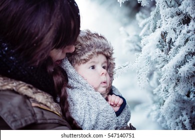 Young Mother Holding Her Newborn Baby With Cap With Ear Flaps. Mother And New Born Boy Having Good Time In The Park In Winter. Baby Boy Is Looking On The Frozen Branch. Color Toned Image.