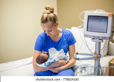 Young Mother Holding Her Premature Newborn Baby Who Is Being Treated In The Hospital. With Love And Tenderness She Holds Her Baby Close While Sitting On A Hospital Bed Next To A Vital Signs Monitor