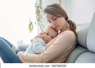 Young Mother, Holding Her Sick Toddler Boy, Hugging Him At Home, Sunny Living Room