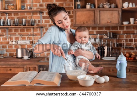 Young mother holding her son, talking on smartphone and mixing a dough at the kitchen. family life and multitasking concept