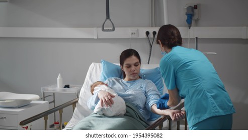 Young Mother Holding Newborn Baby While Nurse Set Catheter In Her Hand. Woman After Childbirth Resting In Hospital Bed With Infant Baby And Medical Worker Adjusting Iv Drip For Her