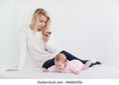 Young Mother Is Holding A Phone. Nearby Lies A Daughter. Mum Is Very Busy And Does Not Pay Attention To The Baby.