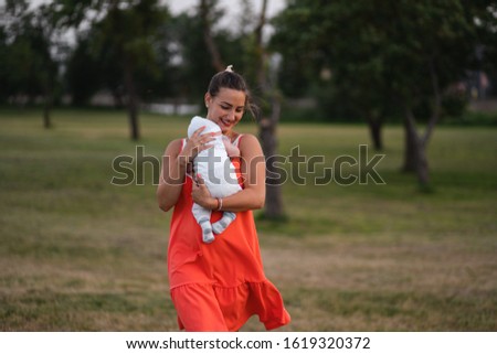 Young mother holding and playing with her baby boy child in city park standing wearing bright red dress - Son wears white cap - Family values warm color summer scene handheld