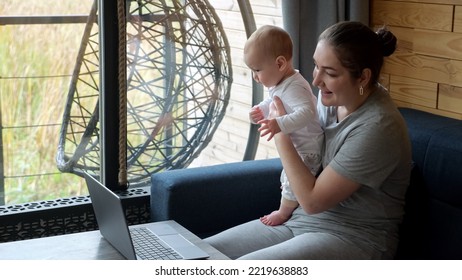 Young Mother Holds Baby Girl On Lap Sitting At Table Near Window. Woman Talks On Video Call Via Laptop Showing Screen To Child With Smile