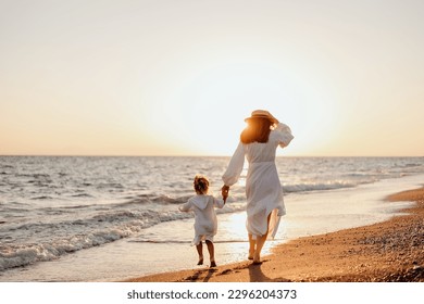 A young mother holds her little daughter by the hand and together they run along the ocean towards the sunset. Girls in white dresses and long hair - Powered by Shutterstock