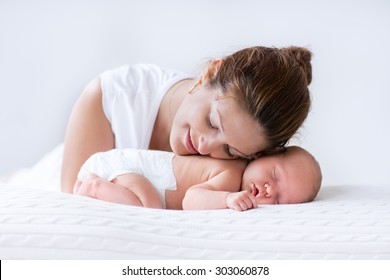 Young mother hugging her newborn child. Mom nursing baby. Woman and new born boy relax in a white bedroom. Family at home. Love, trust and tenderness concept. Bedding and textile for nursery.