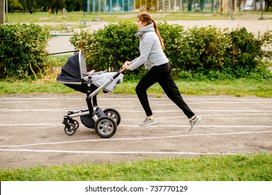 Young Mother Jogging With A Baby Buggy At Stadium Track In Fall Autumn Time. Motherhood, Sport, Run, Fitness Multitasking Concept.