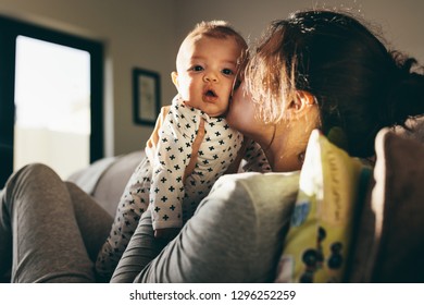 Young Mother Kissing Her Baby Sitting On A Couch At Home. Close Up Of A Woman Holding Her Infant Kid And Pampering Him.