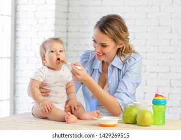 A young mother in the kitchen feeds vegetable puree with a spoon of her 9-month-old child