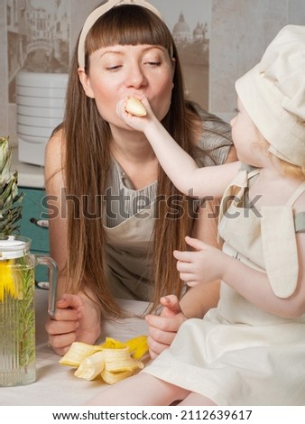Young mother and little daughter eat fruit, banana. Healthy food for children, snack. Caucasian family eats in a light modern kitchen. Daughter feeds mother. Food concept proper. Mother's Day. Diet