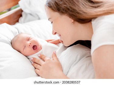 Young mother looking at her sleeping with opened mouth newborn daughter swaddled in white sheets at home and smiling with tenderness. Portrait of girl mom with her infant child napping in the bed - Shutterstock ID 2038280537
