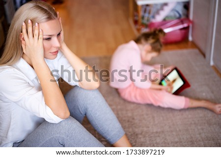 Young mother is nervous and sad at home on the floor, the child watches cartoons on the tablet and plays, naughty child, family concept.