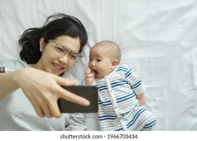 Young Mother And Newborn Baby Lying On Bed Having Fun Taking Selfie Photo With Mobile Phone At Home Together. Asian Mom And Son Enjoying Making Video Call With Their Family On Smartphone