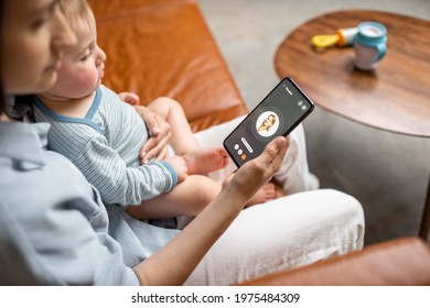 Young Mother With Newborn Kid Making Video Call To A Doctor Using Phone At Home. Concept Of Telemedicine And Support Parents Counseling Online. 