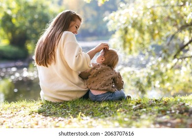 A Young Mother With A One-year-old Baby In An Autumn Park Hugs On The Edge Of The Lake And Smile. Filmed From The Back