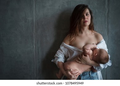 A young mother with a pensive look in white shirt, lowered on her shoulders, and jeans stands at the gray concrete wall, holds the baby in her hands and and breastfeeds.