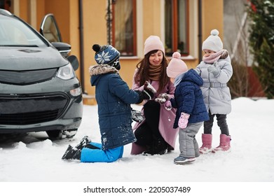 Young Mother Play With Kids Against Electric Car In The Yard Of Her House At Winter.