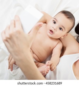 Young Mother Playing And Taking Photo With Smart Phone With Her Baby On White Silk Bed Sheet. Cute Mixed Race Bab Boy Has Fun With Mum, High Key.