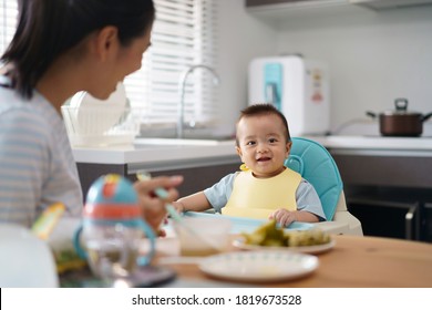 Young Mother Provide Food And Fruit Puree For Lunch To Her Baby Son Seat On High Chair In Kitchen.