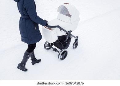 Young Mother Pushing Baby Stroller On Snowy Way. Woman Walking In White Winter Day After Blizzard. Back Side View. 