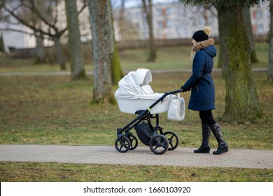 Young mother pushing white baby stroller and slowly walking at town park in cold day. Spending time with infant. Enjoying stroll. City life. Side view. 