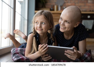 Young Mother Recovering From Oncology And Little Daughter Lying On Pillows On Heated Floor At Warm Comfy Home With Digital Tablet Device In Hands, Looking Outside Through Window Wall Smiling Dreaming