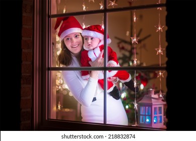 Young Mother In Red Christmas Hat And Her Baby Boy Dressed In Santa Costume Standing Next To A Window In A Decorated Living Room Celebrating Xmas With Lights And Tree. View From Outside Of The House.