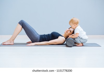 Young Mother Relaxing Her Neck And Back With Foam Roller Does Physical Pilates Exercises While Her Toddler Baby Son Playing Near. Fitness, Happy Maternity Sport With Children Concept.