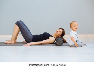 Young Mother Relaxing Her Neck And Back With Foam Roller Does Physical Pilates Exercises While Her Toddler Baby Son Playing Near. Fitness, Happy Maternity Sport With Children Concept.