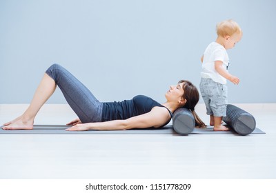 Young Mother Relaxing Her Neck And Back With Foam Roller Does Physical Pilates Exercises While Her Toddler Baby Son Playing Near. Fitness, Happy Maternity Sport With Children Concept.