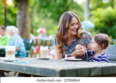 Young Mother Relaxing Together With Her Little Child, Adorable Toddler Girl, In Summer Outdoors Cafe Drinking Coffee And Eating Muffin Or Cupcake.