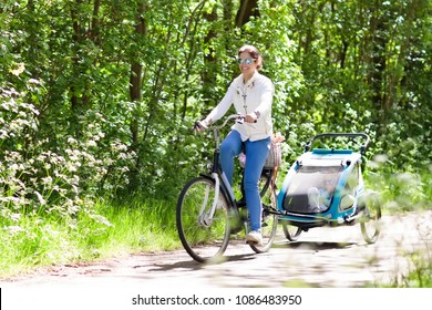 Young Mother Riding Bicycle With Baby Bike Trailer In Sunny Summer Park. Fit Active Woman Cycling With Child. Safe Transportation Of Little Kids. Mom And Children Riding Bikes. Family Outdoor Activity