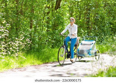 Young mother riding bicycle with baby bike trailer in sunny summer park. Fit active woman cycling with child. Safe transportation of little kids. Mom and children riding bikes. Family outdoor activity