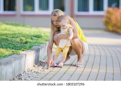 Young Mother Showing Small Stones To Little Daughter During Walk In City In Summer Day. Help To Explore Outdoor Environment.