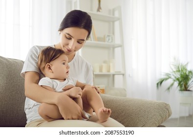 Young mother sitting on sofa together with her little child. Caucasian mom holding her baby son or daughter on her lap while sitting on couch in the living room. Family, care, maternal love concept
