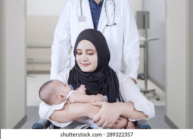 Young Mother Sitting On Wheelchair While Holding Newborn Baby With Her Doctor Pushing The Wheelchair