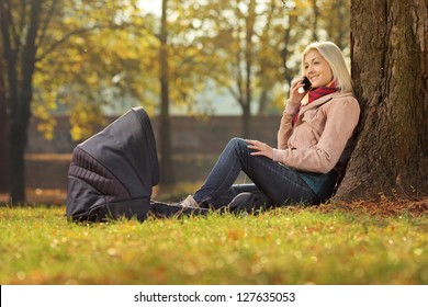 Young Mother Sitting In A Park With Her Baby In A Carrycot And Talking On A Mobile Phone