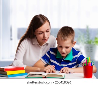Young mother sitting at a table at home helping her small son with his homework from school as he writes notes in a notebook