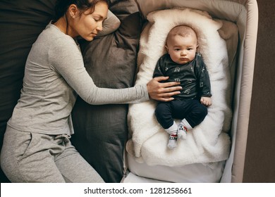 Young Mother Sleeping On Bed With Her Infant Baby Sleeping On A Bedside Crib. Top View Of A Mother Sleeping Beside Her Baby While The Baby Is Awake.