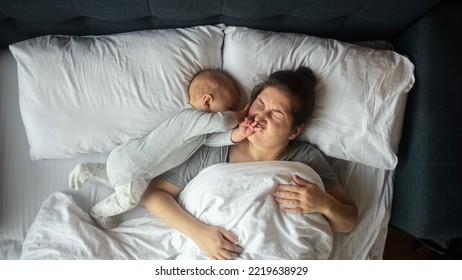 Young mother sleeps with baby girl. Little kid wakes up mother by playing with hair and woman frowns in pain lying on pillows in early morning upper view - Powered by Shutterstock