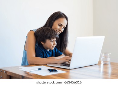 Young Mother And Son Sitting At Table And Using Laptop At Home. Smiling Mom Working At Home With Her Child On The Knees While Writing An Email. Young Woman Teaching Little Boy To Use The Computer.