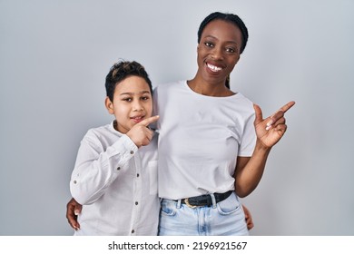 Young Mother And Son Standing Together Over White Background With A Big Smile On Face, Pointing With Hand Finger To The Side Looking At The Camera. 