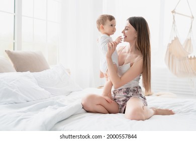 Young mother spending time with her baby son daughter child embracing and hugging kid sitting on the bed. Mom lulling her toddler infant child. Motherhood concept