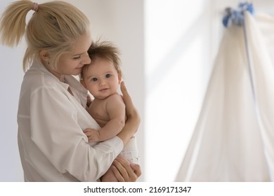 Young mother standing in cozy nursery cuddles, lulls her adorable baby in diaper, copy space. Happy motherhood and maternity, cherish, unconditional love, ad of goods and furniture for infants concept