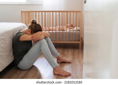 Young mother suffering postnatal depression sitting on floor next to baby napping in bed, resting head on knees, crying, feeling desperate in need of professional psychological support